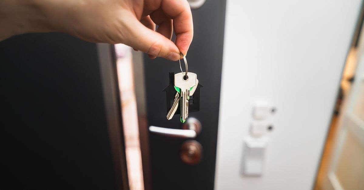 A hand holding a set of house keys on a house-shaped key ring in front of a front door, handing over the keys at the end of a property sale