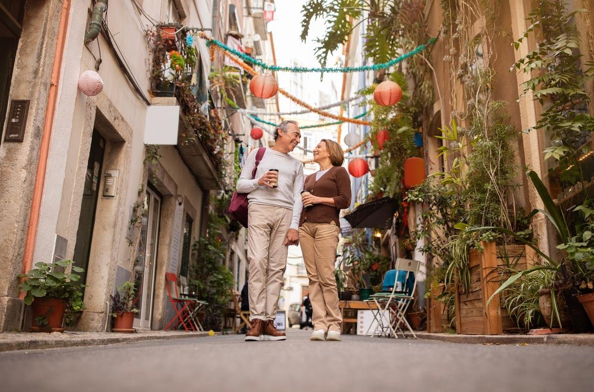A retired couple strolling with coffee cups down a decorated old town lane in Lisbon, Portugal, the retirement move a D7 visa makes possible