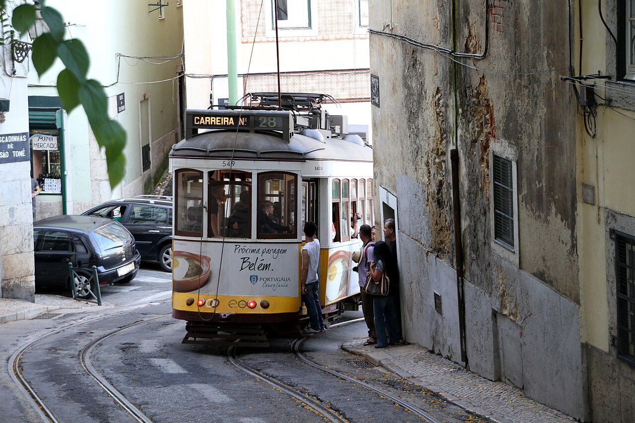 Tram 28 squeezing through a narrow street in Lisbon, Portugal, with passengers boarding, daily life for a family with a Portuguese NIF